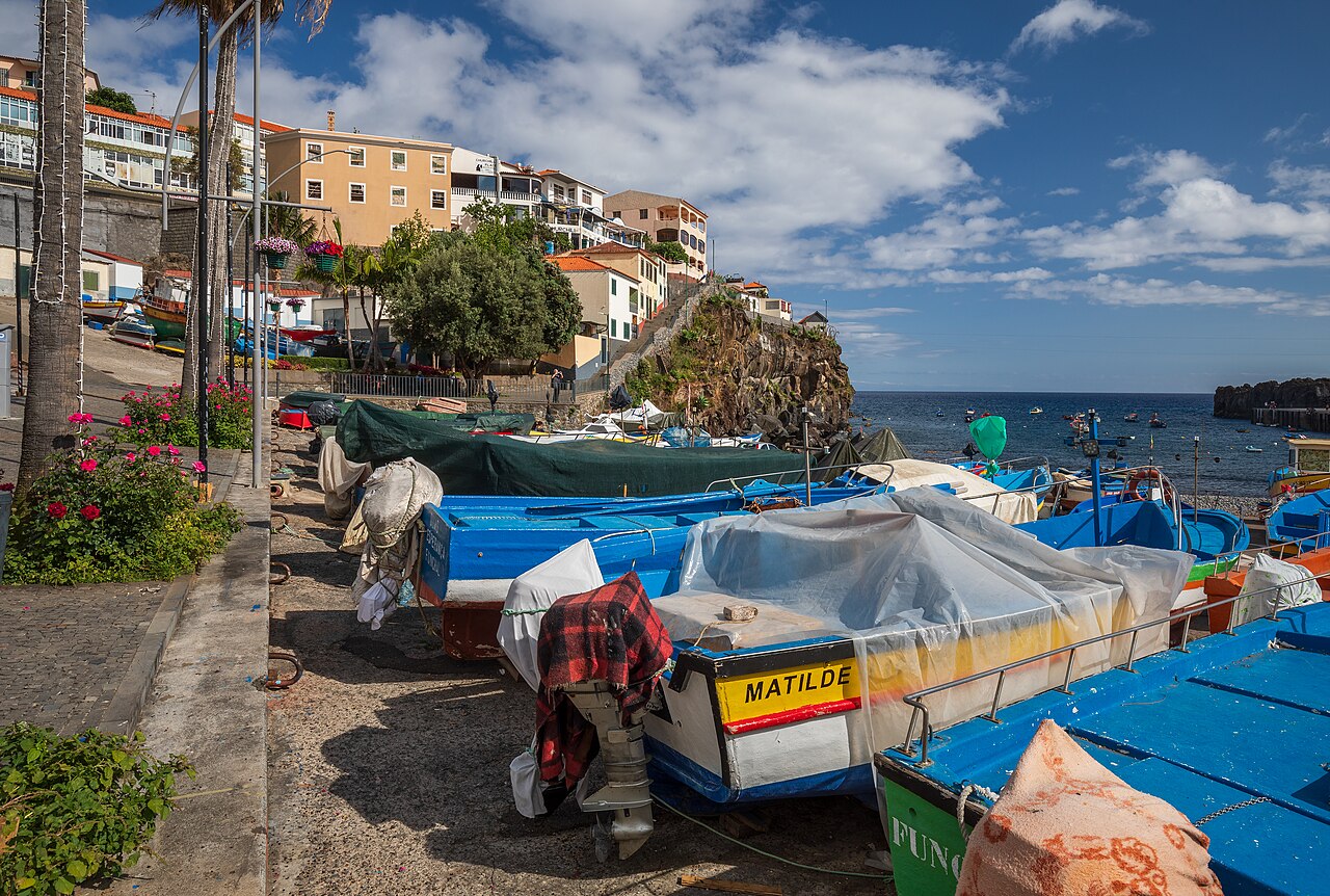 Bunte Fischerboote am Ufer des Dorfes Câmara de Lobos auf Madeira, dahinter Steilküste und Atlantik